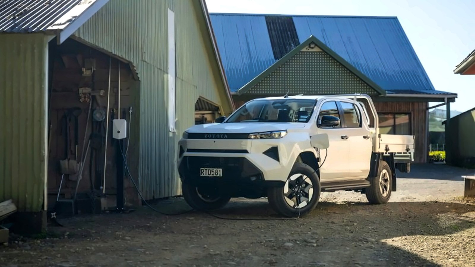 Toyota Hilux BEV cab chassis parked beside a farm shed with wall charger