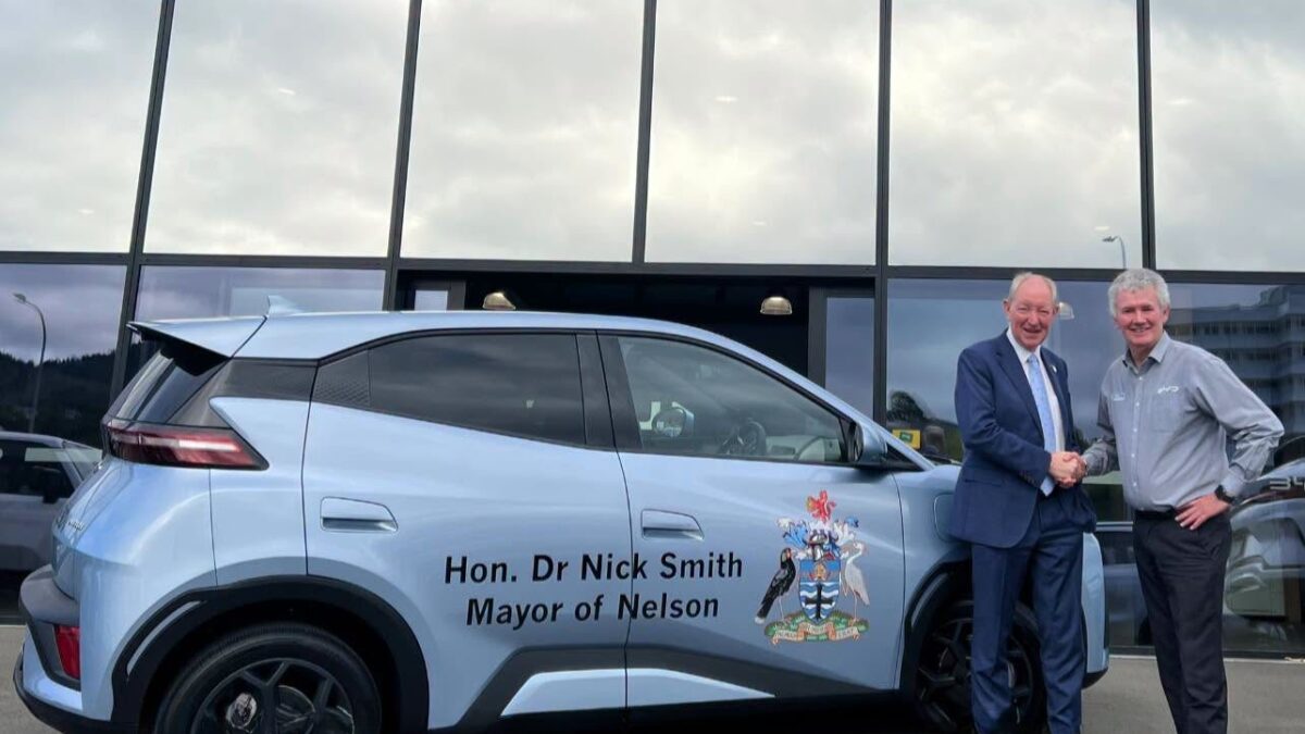 Nelson Mayor Nick Smith shakes hands with The Car Company managing director Shane Drummond beside a light blue BYD Atto 1 with mayoral signwriting outside the BYD showroom in Nelson