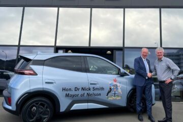 Nelson Mayor Nick Smith shakes hands with The Car Company managing director Shane Drummond beside a light blue BYD Atto 1 with mayoral signwriting outside the BYD showroom in Nelson