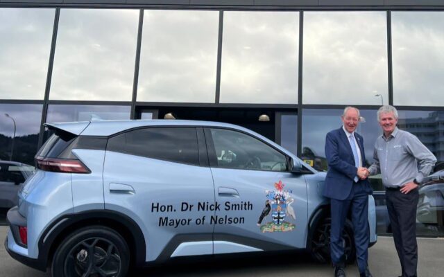 Nelson Mayor Nick Smith shakes hands with The Car Company managing director Shane Drummond beside a light blue BYD Atto 1 with mayoral signwriting outside the BYD showroom in Nelson